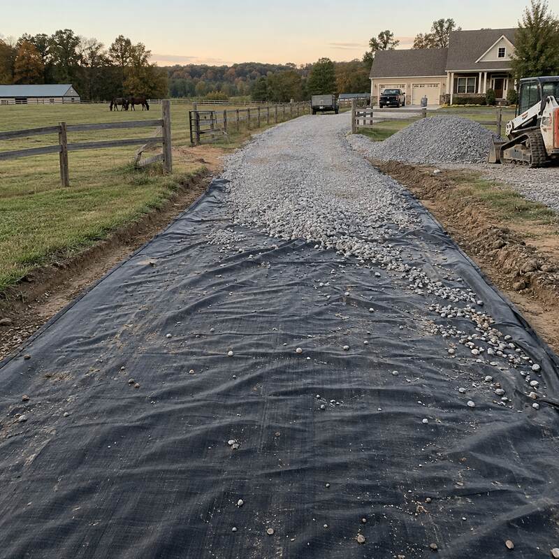 Driveway fabric laid beneath gravel on a horse farm driveway at dusk with horses grazing in the pasture
