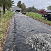 Driveway fabric laid with gravel piled ahead, a tractor and gravel trailer staged near a red barn
