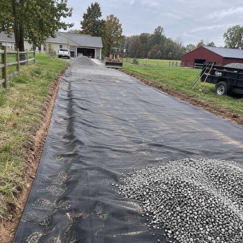 Driveway fabric laid with gravel piled ahead, a tractor and gravel trailer staged near a red barn
