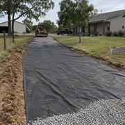 Driveway fabric installed along a rural driveway lined with a stone wall, wheelbarrow and gravel pile nearby
