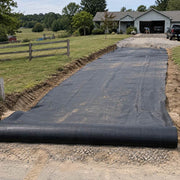 Driveway fabric rolled out over a graded rural driveway leading to a farmhouse with gravel staged ahead
