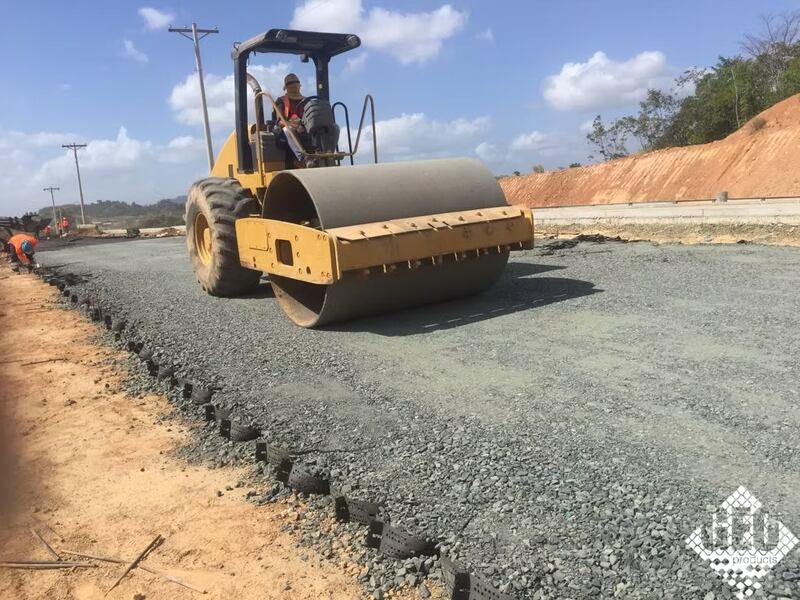 Roller compacting crushed gravel fill over geocell roadway base panels with workers alongside
