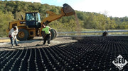 Workers raking gravel over black geocell roadway base panels as a loader dumps additional aggregate
