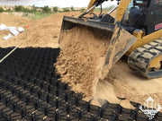 Skid steer loader filling black geocell panels with sand along a desert road base installation
