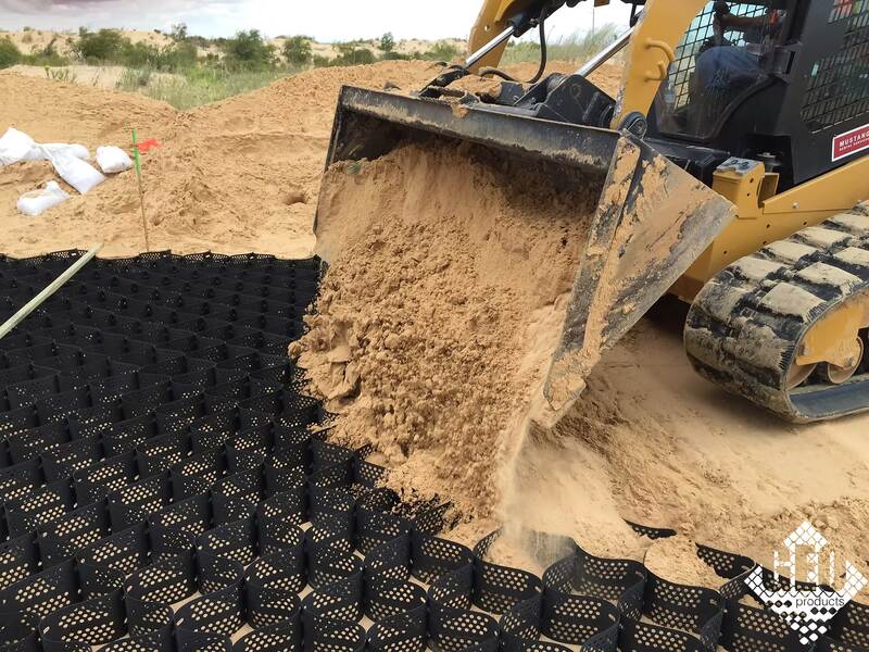 Skid steer loader filling black geocell panels with sand along a desert road base installation
