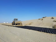 Wheel loader laying a strip of black geocell panels across sandy desert terrain for road base support

