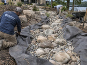 Workers installing non-woven fabric beneath a dry creek bed rock installation at a lakefront home
