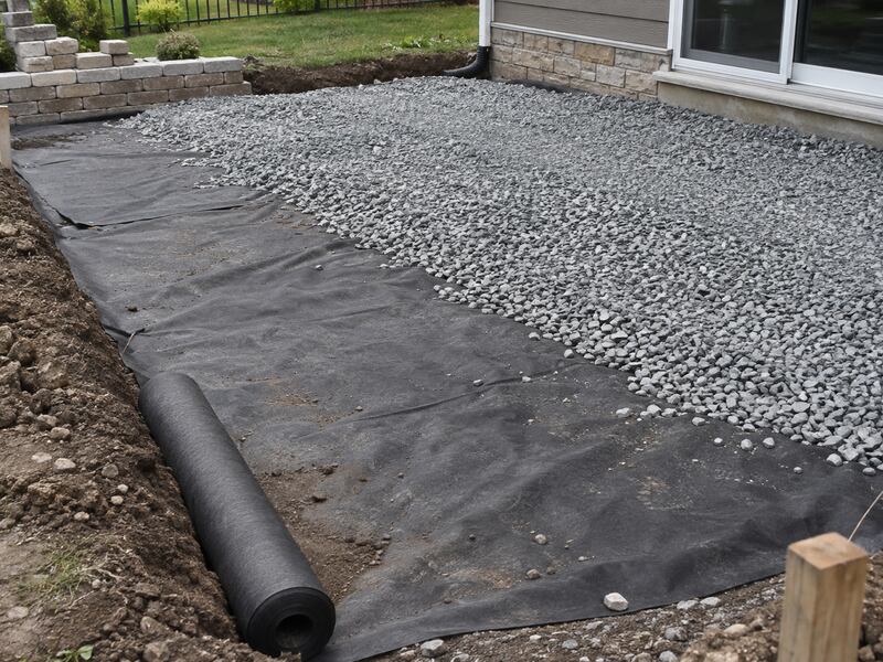 Non-woven fabric laid out with a roll beside a gravel patio base near a house foundation
