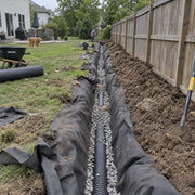 Non-woven geotextile fabric lining a backyard French drain trench with perforated pipe and gravel along a fence line
