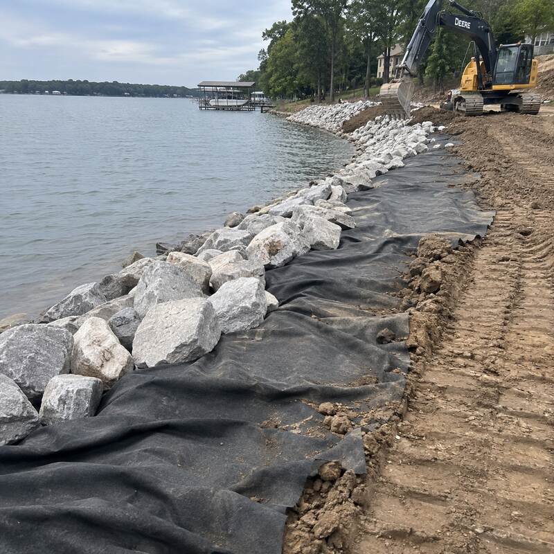 Non-woven geotextile fabric laid beneath rip rap stone along a lakeshore with an excavator placing rock
