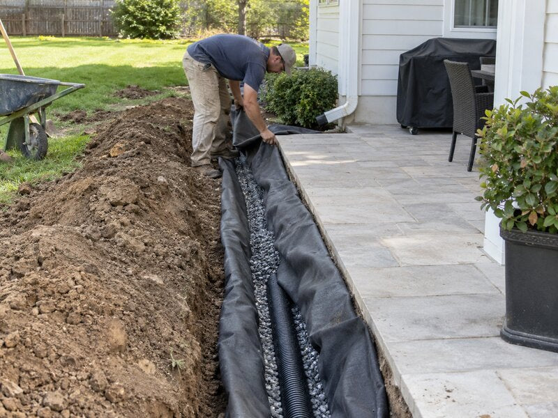 Worker installing non-woven soil filter fabric in a drainage trench beside a paver patio

