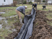 Two men installing non-woven soil filter fabric in a muddy backyard drainage trench
