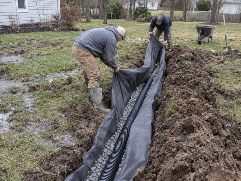 Two men installing non-woven soil filter fabric in a muddy backyard drainage trench
