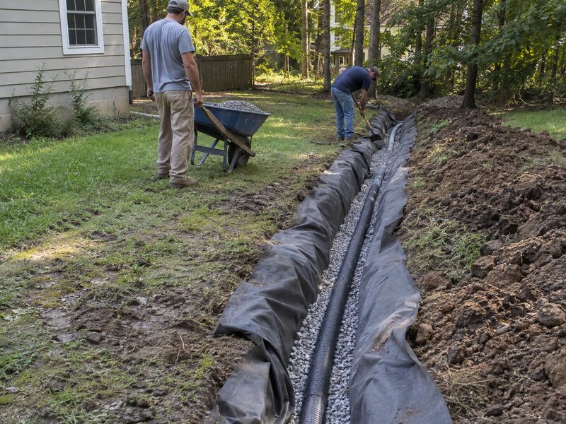 Two men installing non-woven soil filter fabric in a wooded backyard drainage trench with a wheelbarrow
