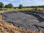 Pond underlayment fabric lining a large farm pond excavation with equipment nearby
