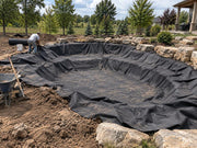 Worker fitting pond underlayment fabric into a stone-edged rural backyard pond excavation

