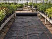 Roll of woven ground cover fabric unrolled down a greenhouse aisle between potted shrubs
