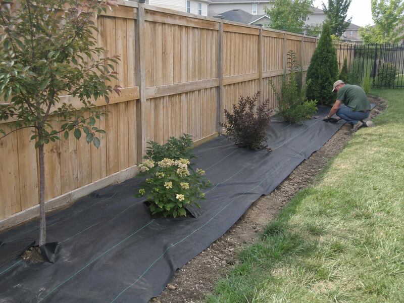 Weed barrier fabric installed along a backyard planting bed beside a wood privacy fence
