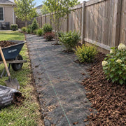 Weed barrier fabric laid along a backyard fence-line planting bed with trees, shrubs, and a mulch wheelbarrow
