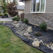 Weed barrier fabric beneath a decorative river rock dry creek bed with boulders along a house foundation
