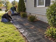 Two women installing woven ground cover fabric along a shrub bed beside a house
